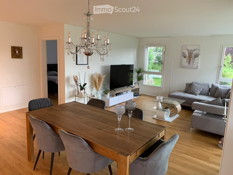 Open-plan living and dining area with a grey sofa, wooden dining table, chandelier, and large windows overlooking a garden.