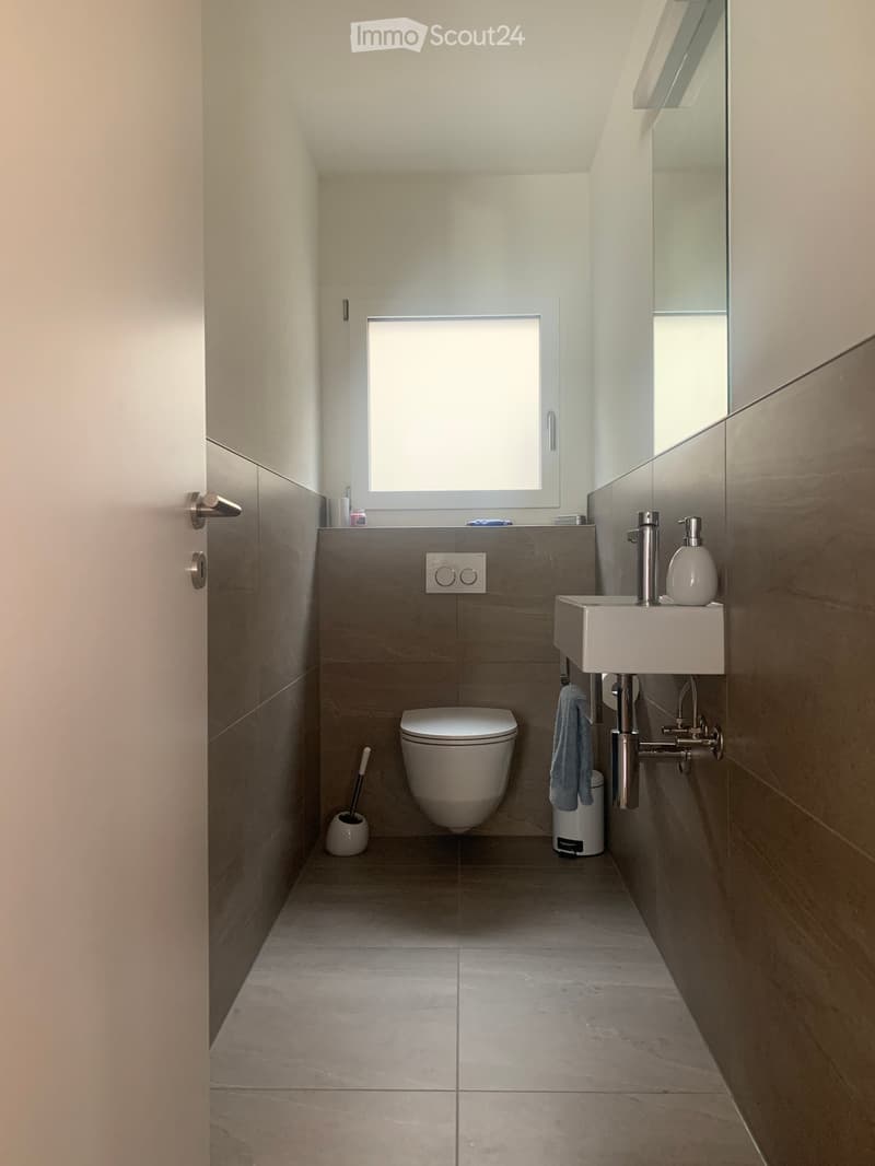 Modern, compact bathroom with a toilet, sink, and mirror. Features include a window, tiled walls, and a towel hanging on the side.