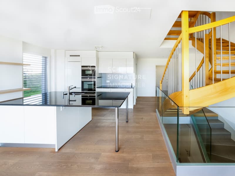 Modern kitchen with white cabinets, stainless steel appliances, a central island with a glass countertop, and a wooden staircase with glass railing leading to an upper level.