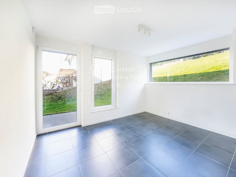 Empty room with white walls, dark grey tiled floor, large windows, and a sliding glass door leading to a green outdoor area.
