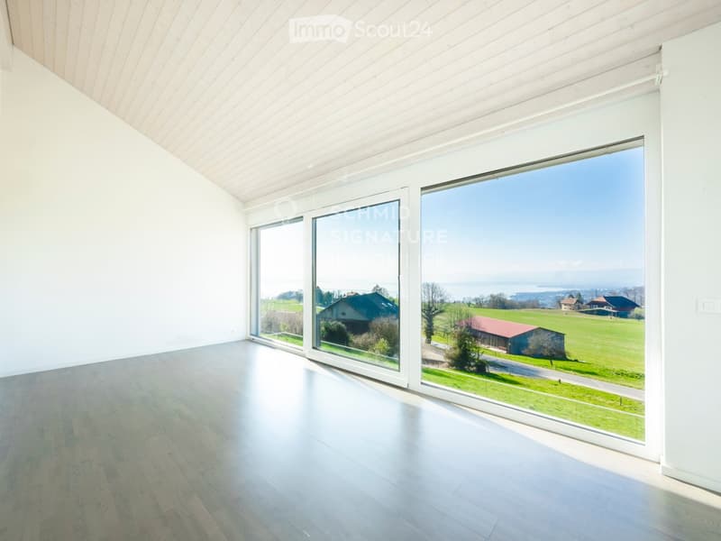 Empty room with white walls, wooden floor, large panoramic windows offering a scenic view of a green landscape and distant houses.