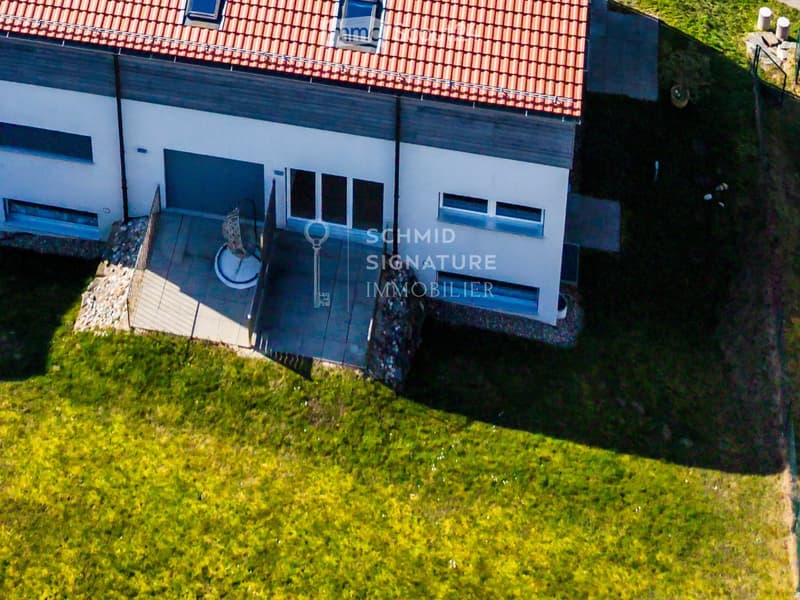 Two-story house with a raised ground floor, red-tiled roof, and modern white facade. The house is situated on a grassy slope with a small patio area visible.