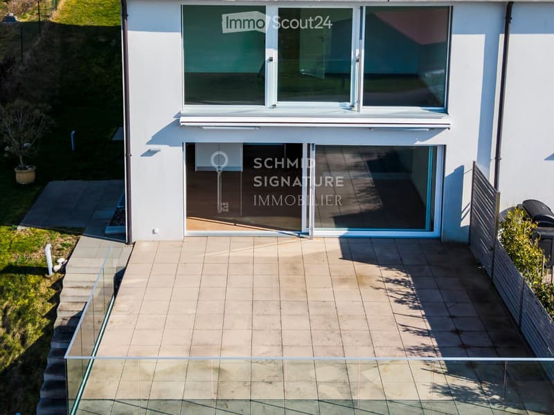 Modern two-story house with large windows, glass doors, and a tiled patio area. The house features a raised ground floor and a landscaped garden with steps leading up to the entrance.