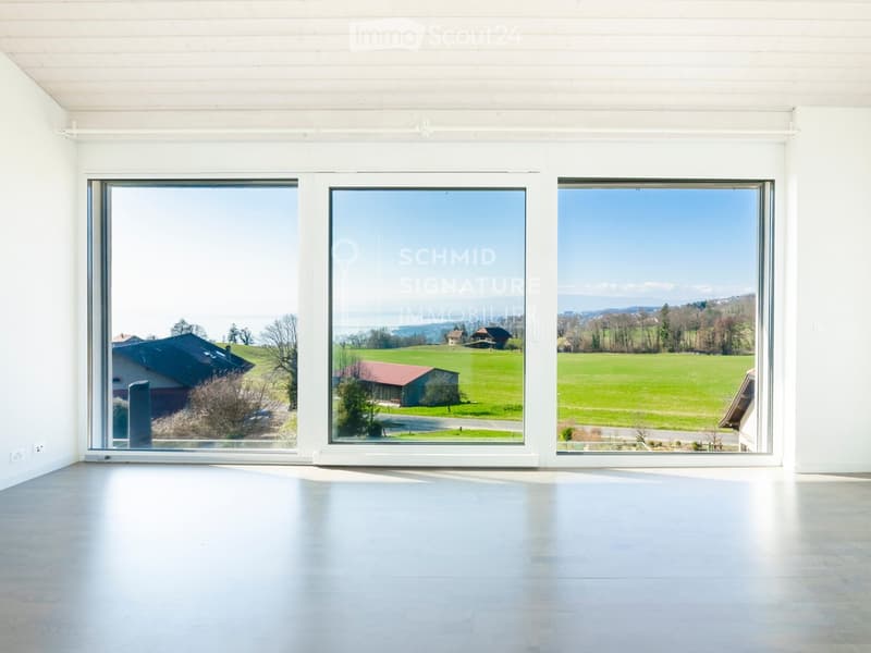 Empty room with white walls, large windows, and a smooth floor, offering a scenic view of a green landscape and houses.
