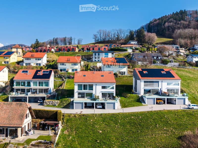Aerial view of a residential neighborhood with multiple modern houses featuring red-tiled roofs, solar panels, and attached garages. The houses are surrounded by green lawns and trees, with a hilly landscape in the background.