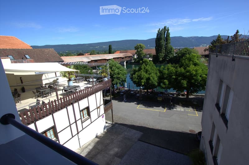 Balcony overlooking a courtyard with outdoor seating, tables, and chairs, surrounded by buildings and trees, with a scenic view of mountains and a river in the background.