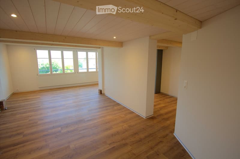 Empty room with wooden flooring, white walls, large windows, and exposed wooden beams on the ceiling.