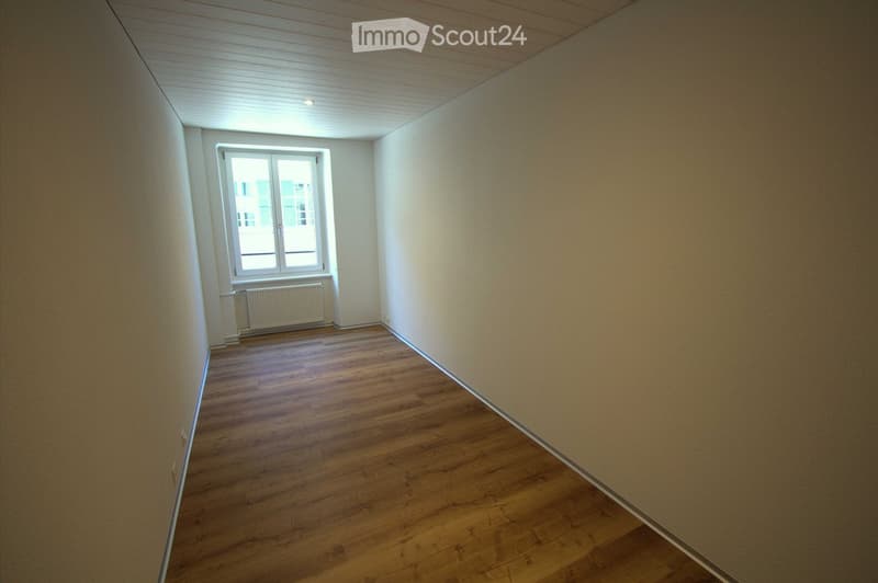 Empty room with wooden flooring, white walls, and a window with a radiator below it.