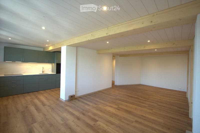 Open-plan space with exposed wooden beams and a modern kitchen area featuring dark green cabinets and stainless steel appliances. The main area has a polished wooden floor and white walls.
