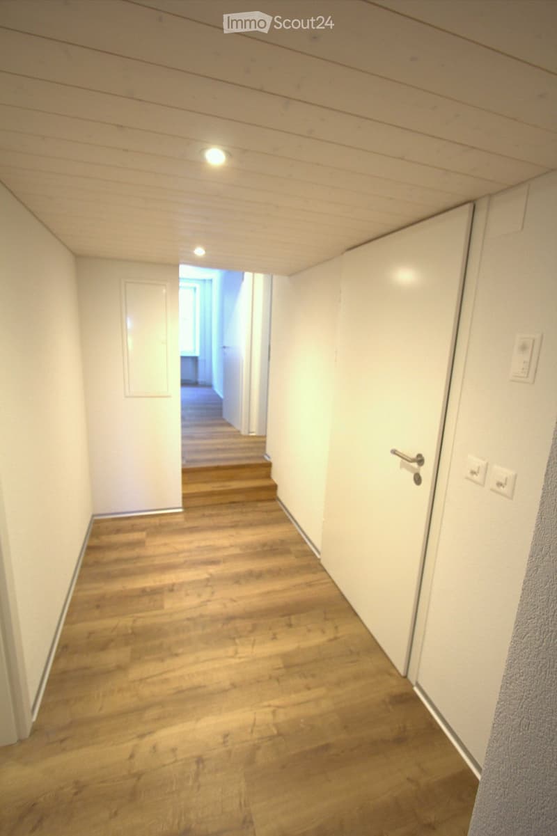 A narrow hallway with wooden flooring, white walls, and recessed lighting. A door is visible on the left, and a staircase leads upward in the background.