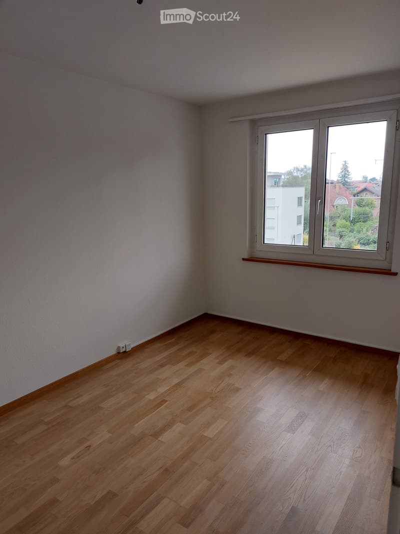 Empty room with light wooden flooring and white walls, featuring a large window with a view of the outside and a small wooden shelf below it.