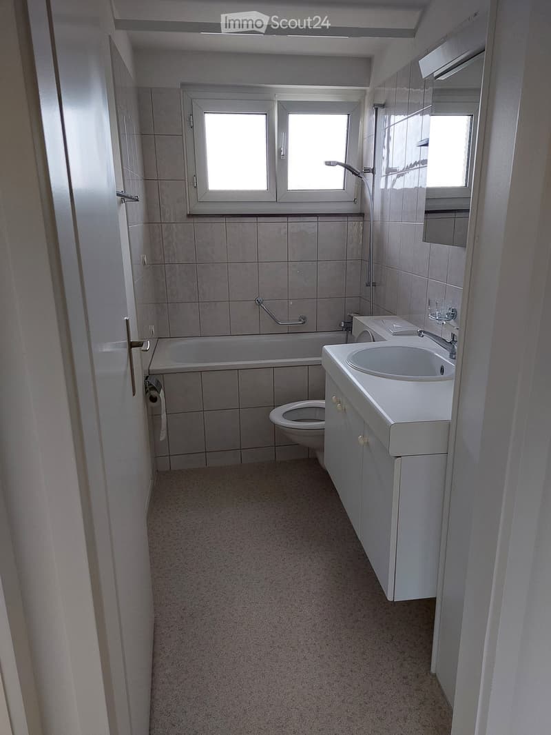 Modern bathroom with tiled walls, bathtub, toilet, sink, and mirror. Features a window and a door with a handle.