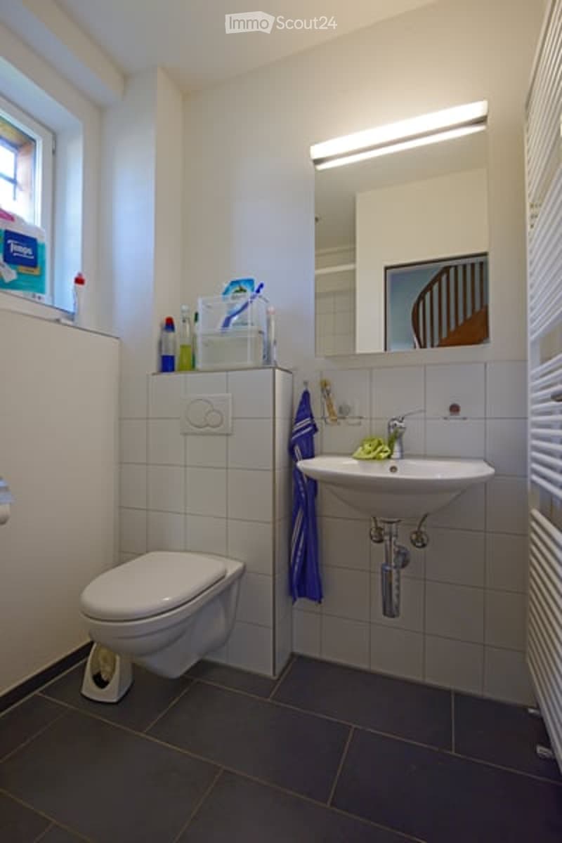 Modern bathroom with white tiles, a toilet, a sink with a mirror above it, and a towel hanging on the side. There are personal care items on the sink counter, including a tissue box, toothbrush holder, toothpaste, shampoo, and shower gel.