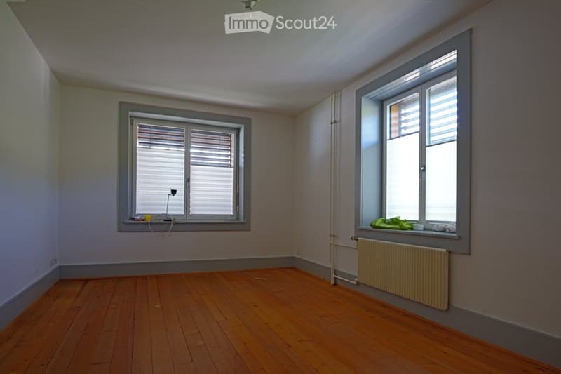 Empty room with wooden flooring, two windows with blinds, radiator, and basic wall fixtures.