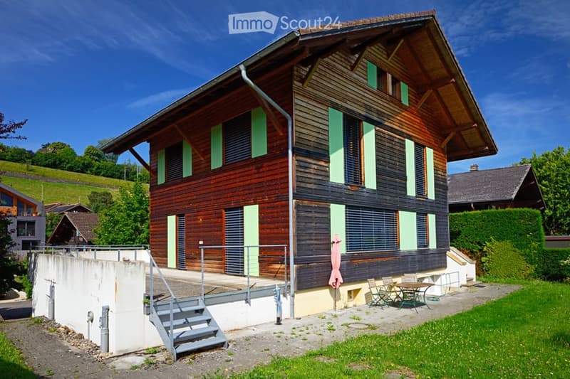 A two-story wooden house with green shutters and a sloped roof, situated in a green, hilly area. The house features a small patio with outdoor furniture, including chairs and a table.