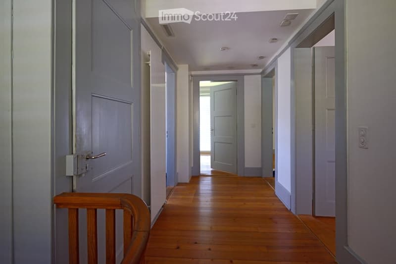A long, narrow hallway with wooden flooring and light blue walls. Multiple doors line the hallway, with one door open to reveal a bright room beyond.