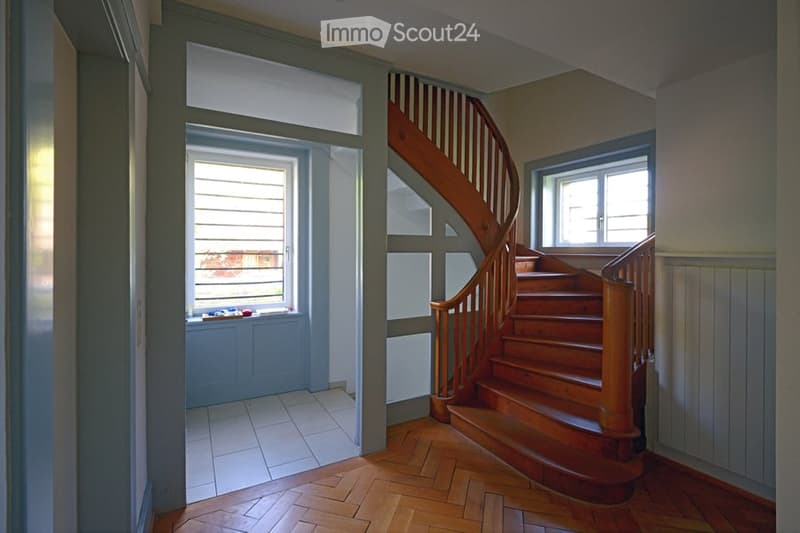 Spacious hallway with wooden flooring, a wooden staircase leading upwards, and large windows providing natural light. The walls are painted in neutral tones, and there is a small window seat with a few decorative items on the left side.