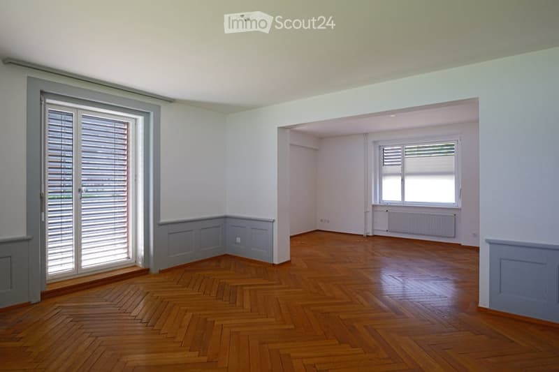 Completely empty room with wooden herringbone flooring, white walls, and large windows with shutters. No furniture or equipment.