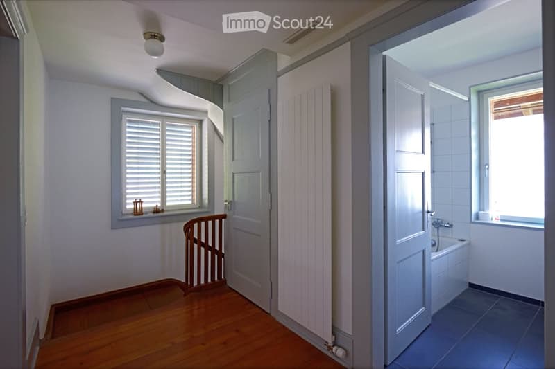 A narrow hallway with wooden flooring, white walls, and a small window with shutters. A door leads to a bathroom with a bathtub and a window, while another door is visible on the opposite side.