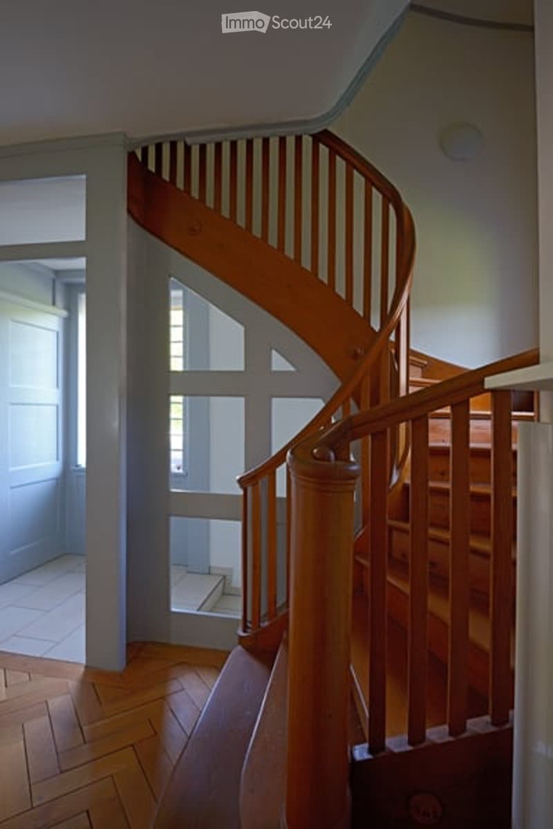 Spiral staircase with wooden handrails and steps, leading to an upper level. The hallway features light-colored walls, wooden flooring, and a door visible in the background.