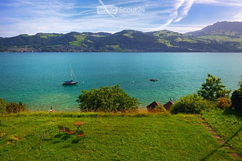 Lush green garden with a scenic view of a lake and mountains, featuring a small table and chairs.