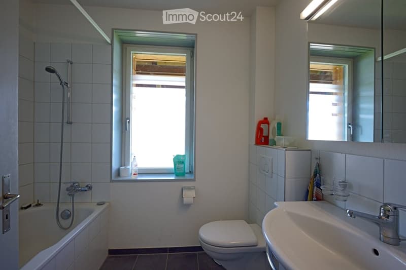 Modern bathroom with a bathtub, toilet, sink, mirror, and a window letting in natural light. The space includes a showerhead, a bottle on the windowsill, and a towel hanging on the side.