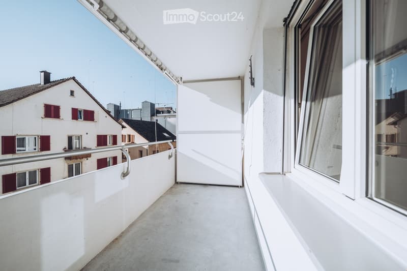 Balcony with white walls, metal railing, and large windows overlooking a cityscape with buildings and rooftops.