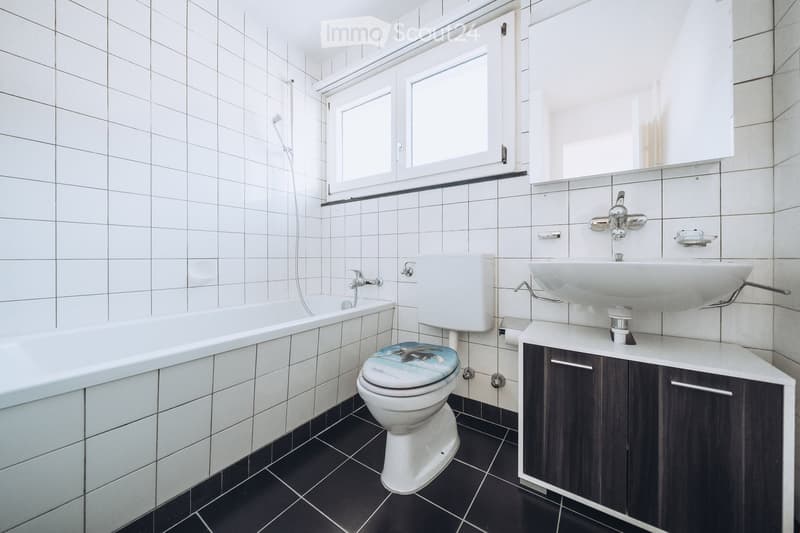 Modern bathroom with white tiled walls, black tiled floor, bathtub, toilet with a decorative seat cover, sink with cabinet, and a window above the bathtub.