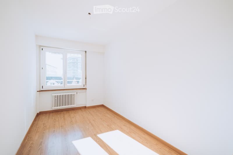 Empty room with white walls, wooden flooring, and a window with a radiator below it.