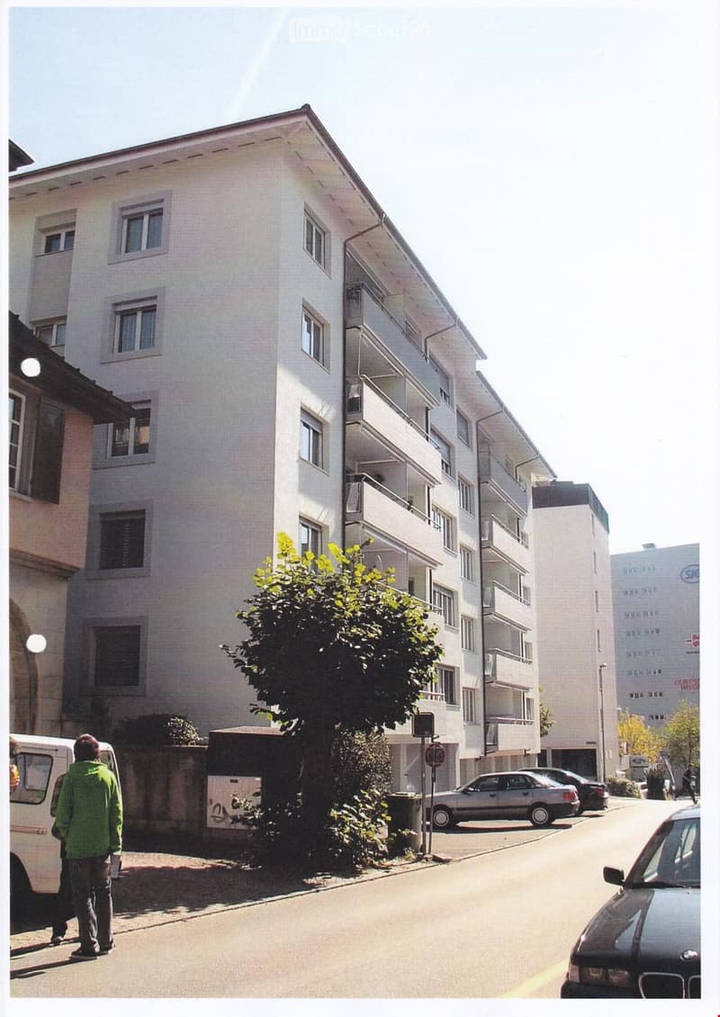 A multi-story residential building with white exterior walls, multiple windows, and balconies. The building is surrounded by parked cars and a tree in the foreground.