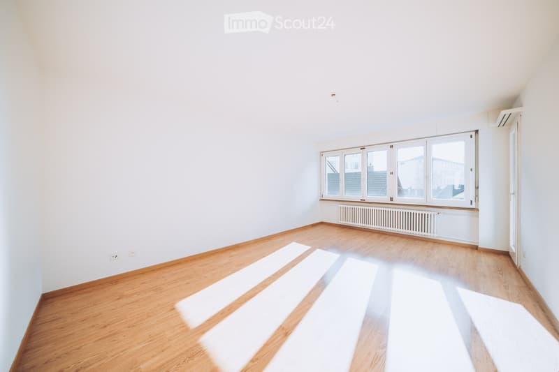 Empty room with wooden flooring and white walls, featuring large windows and a radiator below them.