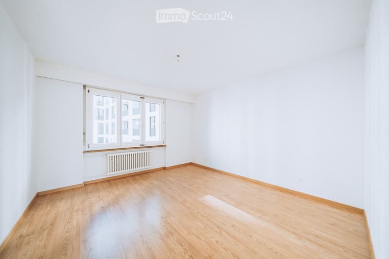 Empty room with white walls, wooden flooring, and large windows with a radiator below. No furniture or equipment present.