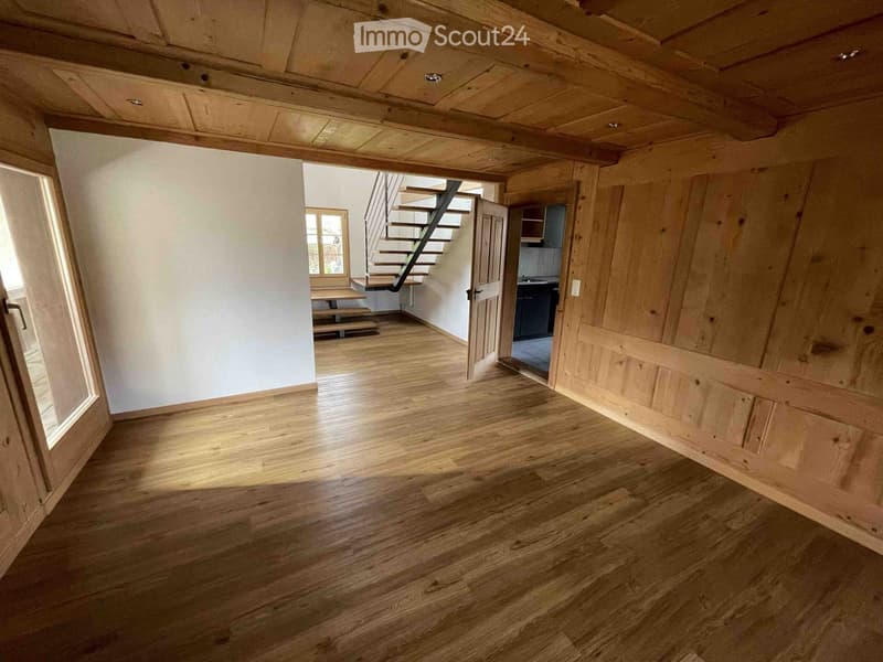 Unfurnished Room. Empty room with wooden flooring, exposed wooden beams on the ceiling, and a staircase leading to another level. The room has white walls and large windows allowing natural light to enter.