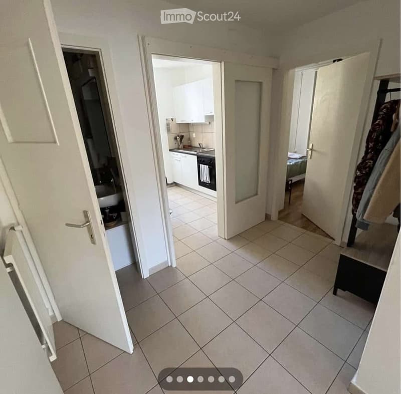 A tiled hallway with white walls, leading to a kitchen and bedroom. The kitchen has white cabinets, a sink, and an oven. The bedroom has a bed and a wardrobe with clothes hanging inside.