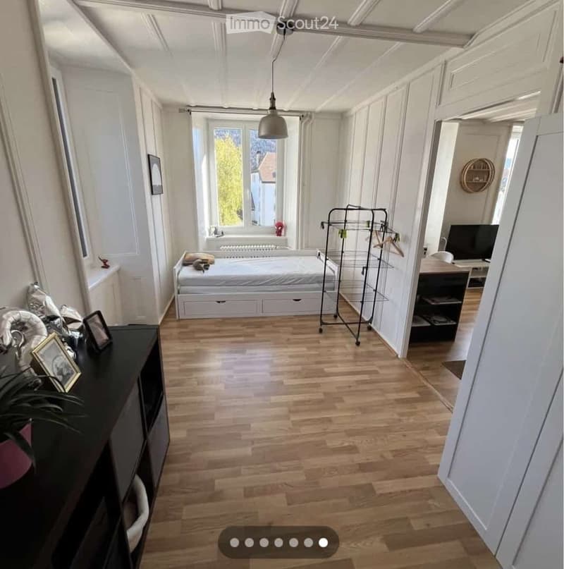 Bright bedroom with wooden flooring, white walls, and large windows. Features a bed with a white mattress, a nightstand, a clothing rack, a desk with a chair, and several picture frames on a black cabinet.