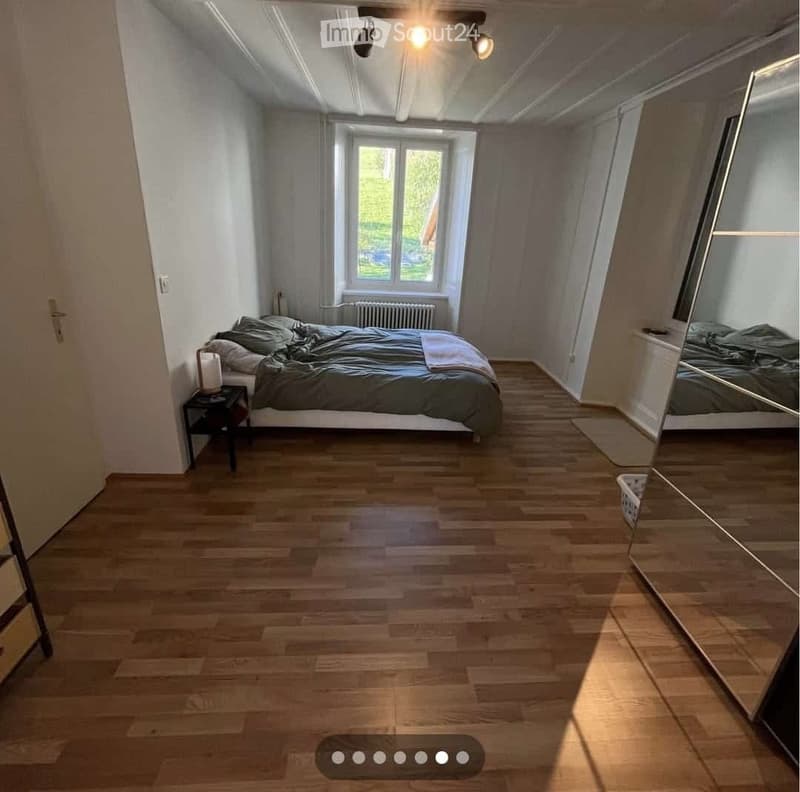 Cozy bedroom with wooden flooring, large window, bed with dark bedding, nightstand, and mirror.