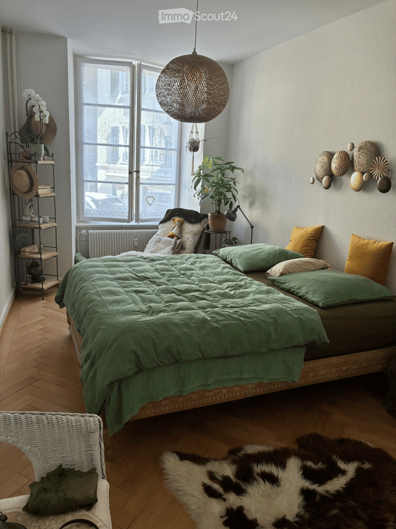 Cozy bedroom with a large bed covered in green bedding, multiple pillows, a wicker hanging light fixture, a wicker chair, a shelf with decorative items, a plant on a side table, and a cowhide rug on the floor.