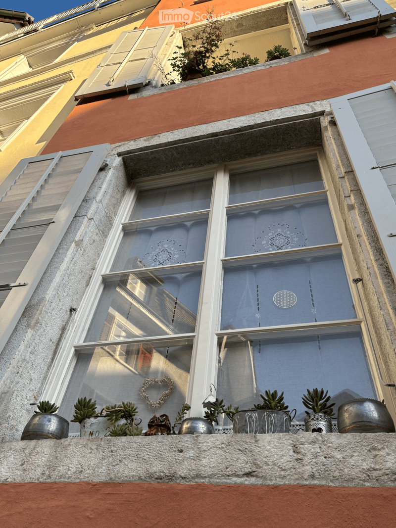 Balcony with multiple potted plants, a heart-shaped decoration, and a window with white shutters and patterned curtains.