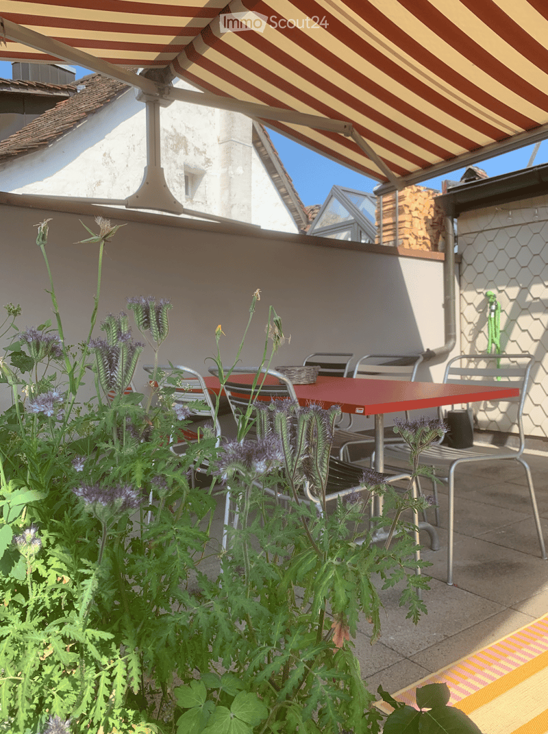Balcony with a red table, several chairs, and lush flowering plants.