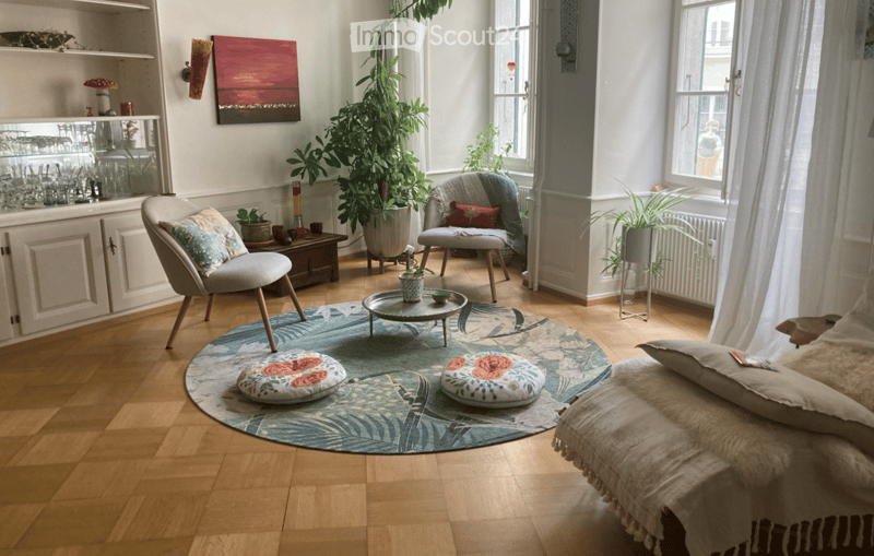 A cozy living room with wooden flooring, a large round rug, a sofa, a chair, a coffee table, and a cabinet with glassware. There are plants and a painting on the wall.