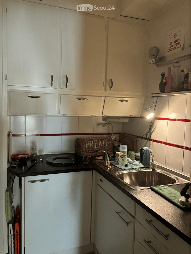 The kitchen features white cabinets with silver handles, a black countertop, and a stainless steel sink. A wicker basket labeled "BREAD" sits on the counter along with various kitchen items including cups, bottles, and a green towel.