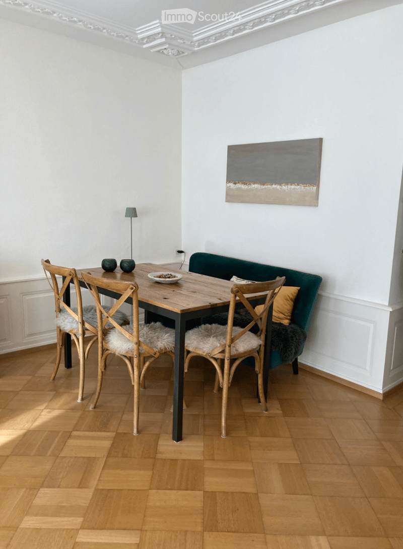 A modern dining area with a wooden table, four chairs with fur cushions, a green sofa with a yellow pillow, a small lamp, and a contemporary painting on the wall.