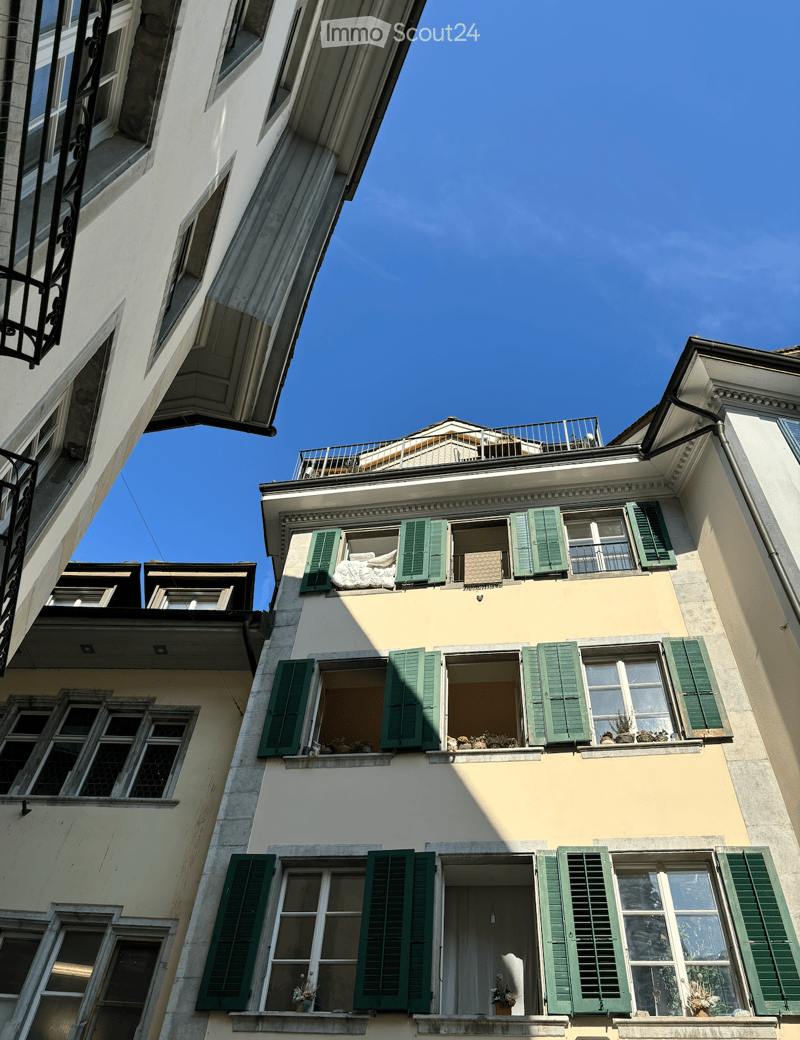 Multi-story residential building with green shutters, windows, and balconies. Visible details include a rooftop terrace and various window decorations.