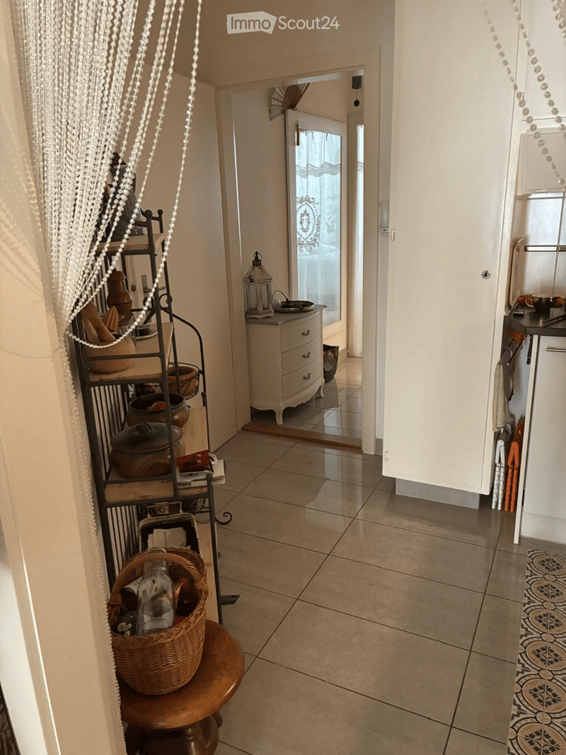 A hallway with tiled flooring, a small wooden stool with a woven basket, a metal shelf with decorative items, and a small white cabinet with drawers.