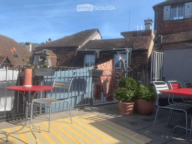 Balcony with red tables, chairs, potted plants, and a striped rug.