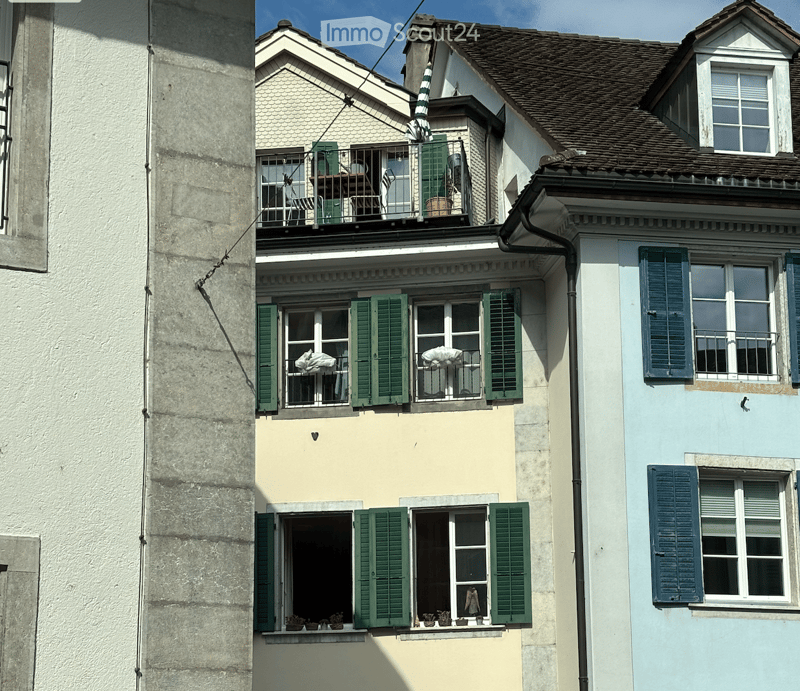 Multi-story building with green shutters, balconies, and potted plants on the windowsills.