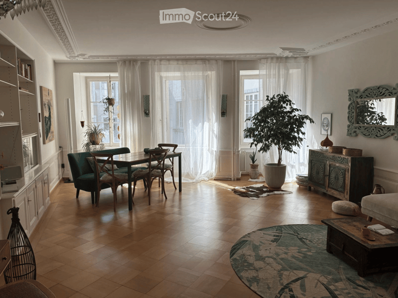 Bright living room with wooden flooring, large windows with sheer curtains, a green sofa, coffee table, dining table with chairs, a large potted plant, and decorative cabinet.