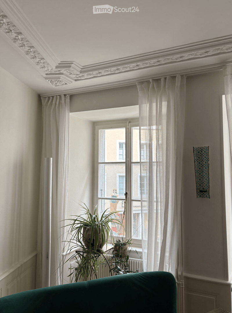 A well-lit room with a green sofa, large window with white curtains, and potted plants on the windowsill.
