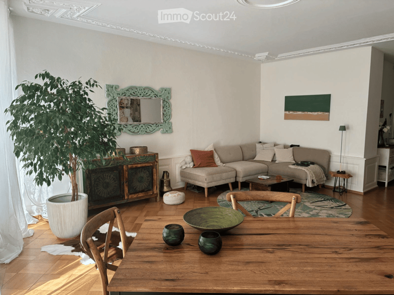 Living room with a sectional sofa, coffee table, wooden dining table, chairs, a large potted plant, a decorative cabinet, and artwork on the walls.