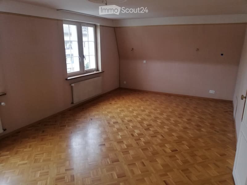 Empty room with wooden parquet flooring, light-colored walls, and a large window with a radiator below it.